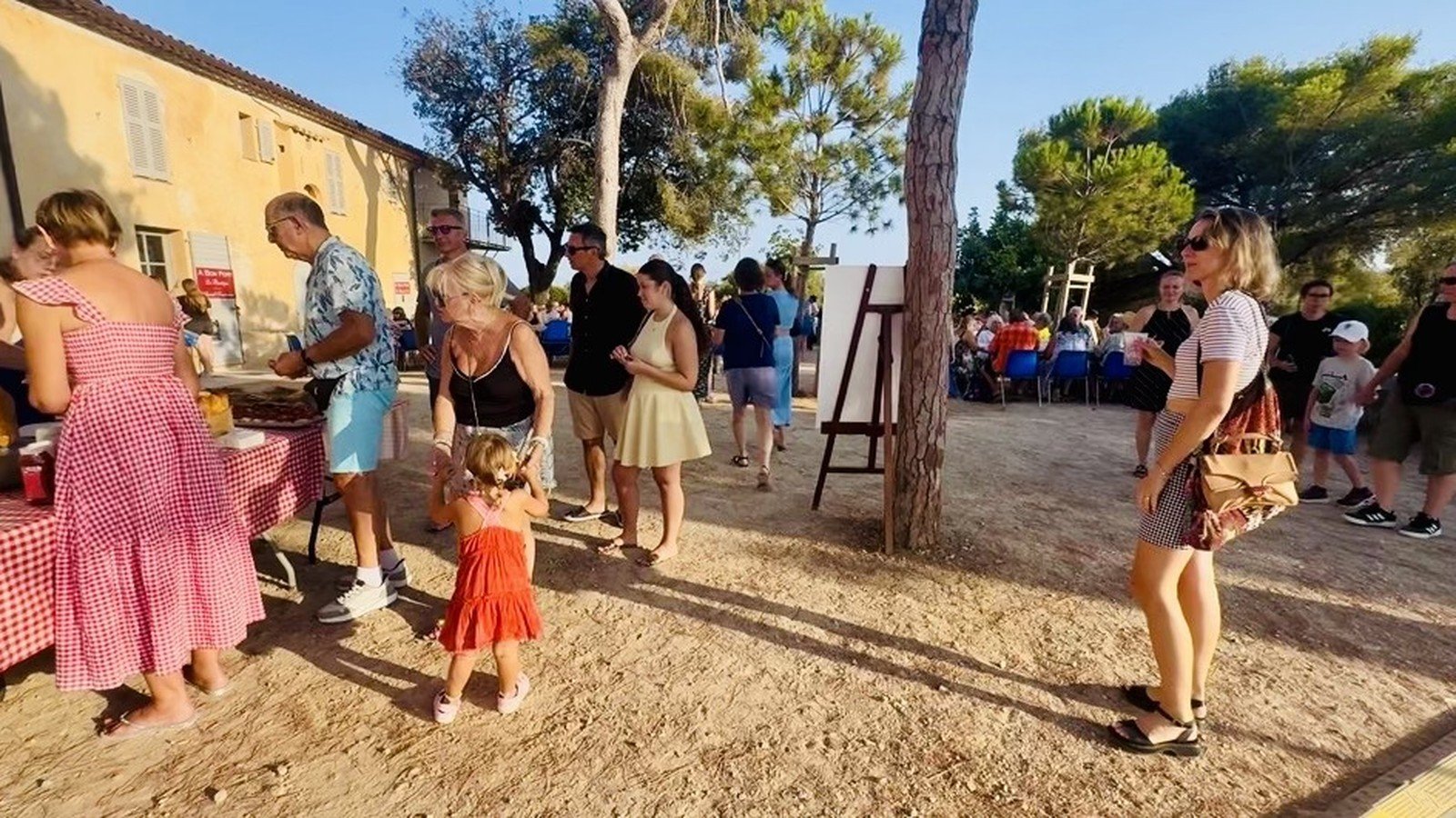 Une fête de village en fin de journée sous les pins, des familles autour d’un stand et des tables installées en plein air