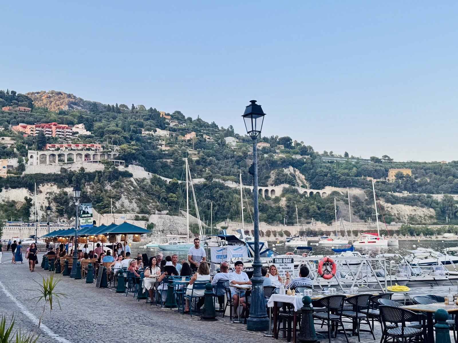 Restaurant tables along the Villefranche-sur-Mer harbor in the evening, boats anchored in the bay