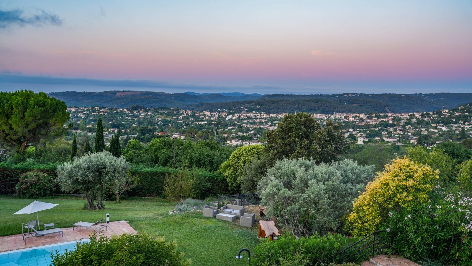 A villa garden terrace with a swimming pool and olive trees, overlooking a hillside village and valley at dusk